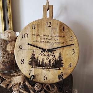 May include: A round wooden clock with black hands and numbers. The clock face features the text "He who dwells in the shelter of the Most High will abide in the shadow of the Almighty" and a forest silhouette. A jute rope is attached for hanging.