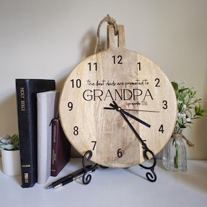 May include: A round wooden wall clock with a handle and jute rope hanger. The clock face features black numbers 1 through 12, black hands, and the text: The best dads are promoted to GRANDPA proverbs 17:6. It rests on a black metal display stand.