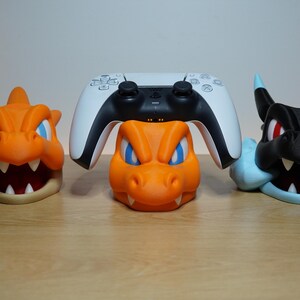 May include: Three 3D-printed controller stands shaped like dragon heads. Two are orange with blue eyes and white teeth, while the third is black with red eyes and light blue accents. A white and black game controller rests on the center stand.