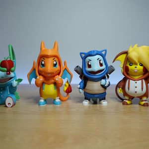 May include: Four small collectible figurines standing in a row. The first is a blue creature in a hooded outfit holding a red apple. The second is an orange dragon-like figure. The third is a blue turtle-like character. The fourth is a yellow creature with spiky hair wearing a brown hooded onesie.