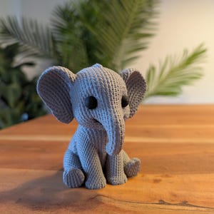 May include: A handmade gray crochet elephant figurine with black button eyes and a long trunk. The elephant is sitting on a wooden surface, with green foliage in the background. The elephant is approximately 6 inches tall.