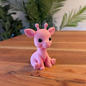 May include: A handmade, pink crocheted giraffe figurine with black safety eyes. The giraffe is sitting with its legs folded and has small ears and horns. The toy is made of a soft, textured yarn.