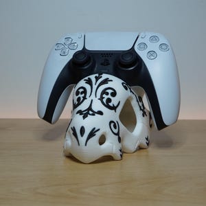 May include: A white and black wireless video game controller resting on a white stand with a black filigree pattern. The controller features a central touchpad, two analog sticks, a directional pad, and four action buttons on the right side.