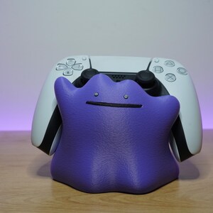 Könnte beinhalten: Ein violetter, amorpher, klecksförmiger Controller-Ständer mit zwei kleinen kreisförmigen Augen und einer dünnen horizontalen Linie als Mund, der einen weißen und schwarzen kabellosen Videospiel-Controller mit texturierter Oberfläche hält.