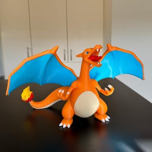 Könnte beinhalten: Orangefarbene Charizard-Figur mit blauen Flügeln und einer gelb-roten Flamme am Schwanz. Die Drachenfigur hat einen offenen Mund mit roter Zunge und weißen Krallen. Die Figur steht auf einer schwarzen Oberfläche.