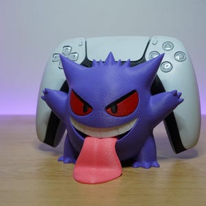 May include: A purple Gengar character figure with red eyes and a long pink tongue, serving as a stand for a white and black video game controller. The figure has a textured surface and is positioned in front of the controller on a flat surface.