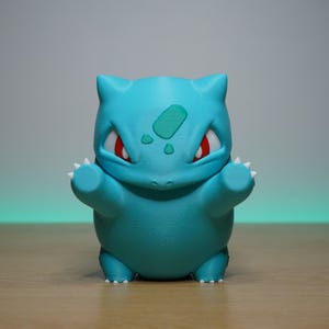 Puede incluir: Figura de Bulbasaur turquesa con ojos rojos y garras blancas. La figura tiene un parche verde en la frente y posa con los brazos extendidos. El fondo es un degradado de gris y verde.