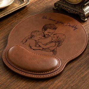 May include: A brown leather mouse pad with a wrist rest featuring an engraved illustration of a parent hugging two children. The text "We love you, Daddy" is engraved in a cursive script above the illustration. The mouse pad has stitched edges.
