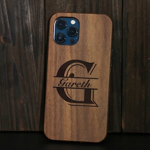 Puede incluir: Funda de teléfono de madera con un diseño personalizado. La funda es de madera de color marrón claro y presenta el nombre "Gareth" grabado en una fuente estilizada, con la letra "G" prominentemente mostrada encima del nombre. Las lentes de la cámara del teléfono son visibles.