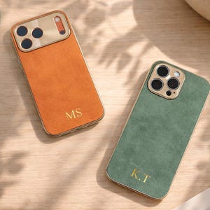 Może przedstawiać: Dwa spersonalizowane etui na telefon. Jedno jest pomarańczowe ze złotymi obramowaniami i inicjałami "MS". Drugie jest zielone ze złotymi obramowaniami i inicjałami "K.T". Oba etui mają złotą osłonę obiektywu aparatu.