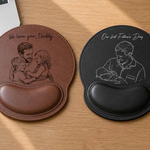 Op de afbeelding: Twee ronde muismatten met polssteunen, één bruin en één zwart. De bruine mat heeft een gegraveerde illustratie van een ouder met twee kinderen en de tekst "We love you, Daddy". De zwarte mat toont een gegraveerde illustratie van een ouder met een baby en de tekst "Our first Father's Day".