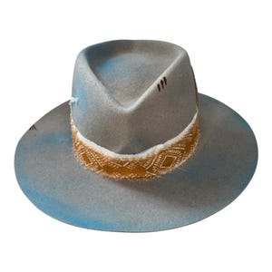 Può includere: Un cappello grigio in stile fedora con tesa larga e fascia decorativa. La fascia è color cuoio con un motivo geometrico bianco. Il cappello ha un aspetto invecchiato con accenti blu sbiaditi sulla tesa e sulla corona. Sono visibili piccoli segni neri.