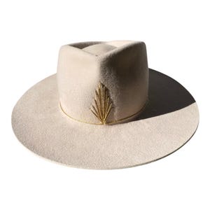 Könnte beinhalten: Ein cremefarbener Fedora-Hut mit breiter Krempe. Der Hut hat ein goldfarbenes, gesticktes Blattdesign auf der Vorderseite und ein goldfarbenes Band um die Basis des Hutes. Der Hut ist aus einem weichen, filzartigen Material gefertigt.