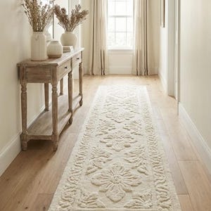 Puede incluir: Un pasillo con una alfombra crema con estampado floral sobre un suelo de madera clara. La alfombra conduce a una ventana con cortinas crema. A la izquierda hay una consola de madera con jarrones y plantas secas. El dise&ntilde;o de la alfombra es visible.