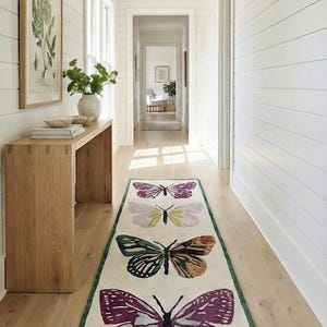 May include: A hallway runner rug with four butterfly designs in shades of purple, green, and brown on an off-white background. The rug has a green border and is placed on a light wood floor.