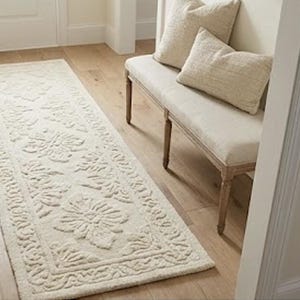 Puede incluir: Alfombra rectangular color crema con un patr&oacute;n floral, colocada sobre un suelo de madera. La alfombra presenta un dise&ntilde;o detallado con motivos florales en relieve y un borde decorativo. Un banco con dos almohadas es visible.