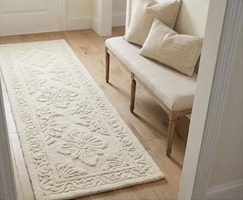 Puede incluir: Alfombra rectangular color crema con un patr&oacute;n floral, colocada sobre un suelo de madera. La alfombra presenta un dise&ntilde;o detallado con motivos florales en relieve y un borde decorativo. Un banco con dos almohadas es visible.