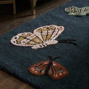 May include: A dark teal rug featuring three embroidered butterfly designs. The butterflies have detailed wings in shades of pink, gold, brown, and green. The rug is placed on a dark wood floor, with a glimpse of a sofa and gold table leg.