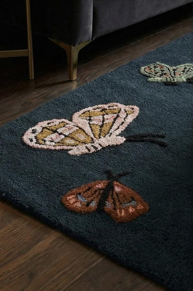 May include: A dark teal rug featuring three embroidered butterfly designs. The butterflies have detailed wings in shades of pink, gold, brown, and green. The rug is placed on a dark wood floor, with a glimpse of a sofa and gold table leg.