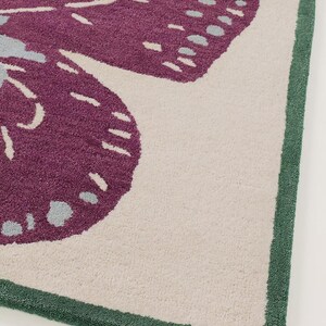 May include: Close-up of a rug featuring a large butterfly design. The butterfly has purple wings with light blue and white accents, set against a cream-coloured background. The rug is bordered with a green trim.