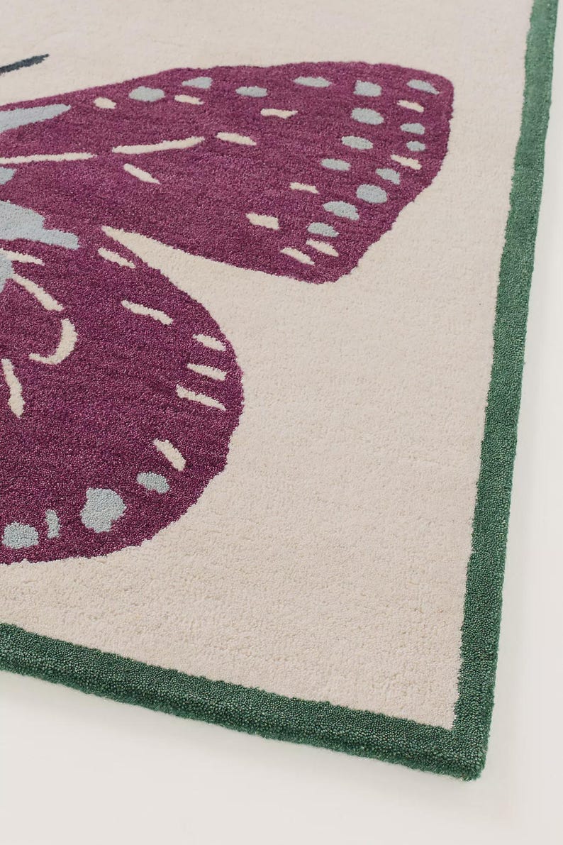 May include: Close-up of a rug featuring a large butterfly design. The butterfly has purple wings with light blue and white accents, set against a cream-coloured background. The rug is bordered with a green trim.
