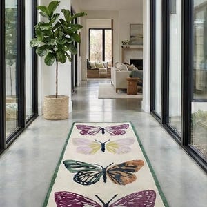 May include: A rectangular rug featuring four butterfly designs in shades of purple, green, and orange on an off-white background. The rug has a green border and is placed in a hallway with large windows.