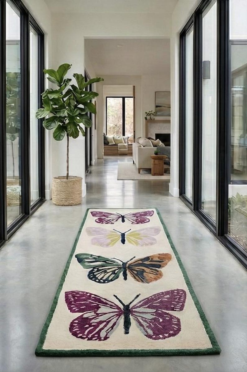 May include: A rectangular rug featuring four butterfly designs in shades of purple, green, and orange on an off-white background. The rug has a green border and is placed in a hallway with large windows.