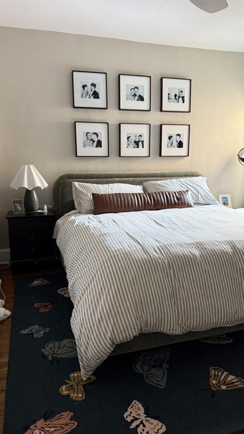 May include: A bedroom scene featuring a bed with a striped comforter and a brown leather bolster pillow. Above the bed, six framed black and white photos are displayed. A dark rug with butterfly designs is on the floor.
