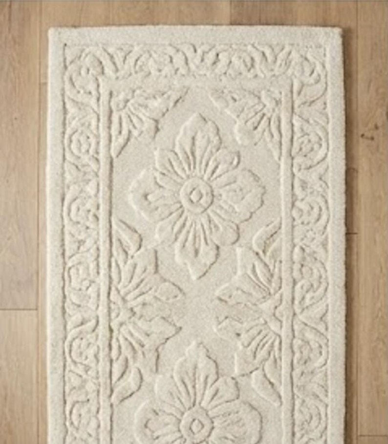 Puede incluir: Alfombra de ba&ntilde;o rectangular de color crema con un estampado floral. La alfombra presenta un dise&ntilde;o en relieve texturizado de grandes flores y hojas, enmarcado por un borde decorativo. El color neutro y el patr&oacute;n detallado crean un aspecto cl&aacute;sico y elegante.
