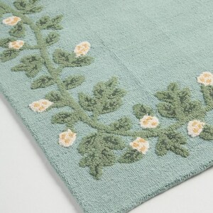 May include: A rectangular, pale teal rug with a floral pattern. The rug has a border of green leaves and white and yellow flowers. The design is embroidered, giving a textured feel. The rug is likely made of cotton.