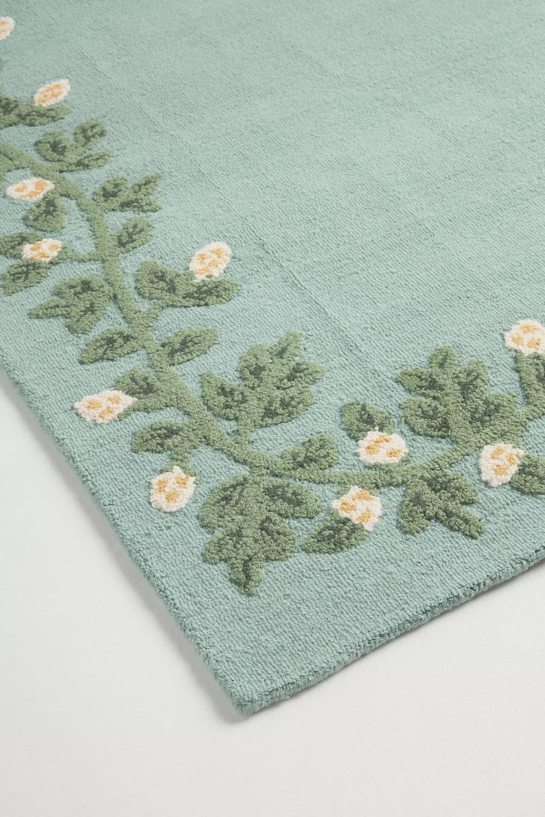 May include: A rectangular, pale teal rug with a floral pattern. The rug has a border of green leaves and white and yellow flowers. The design is embroidered, giving a textured feel. The rug is likely made of cotton.