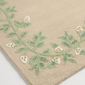 May include: Beige rug with a green and white floral pattern. The rug has a repeating design of green leaves and white flower buds. The design is embroidered, creating a textured effect on the rug's surface.