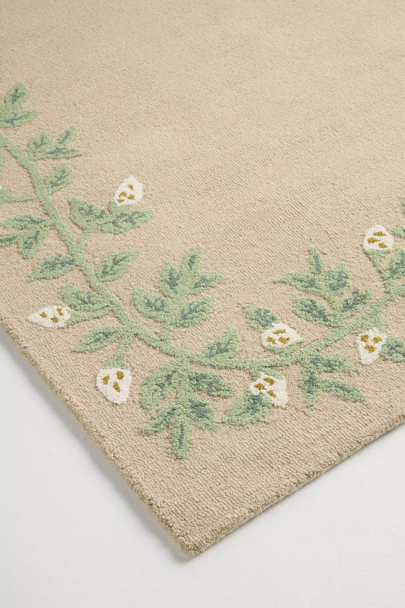 May include: Beige rug with a green and white floral pattern. The rug has a repeating design of green leaves and white flower buds. The design is embroidered, creating a textured effect on the rug's surface.