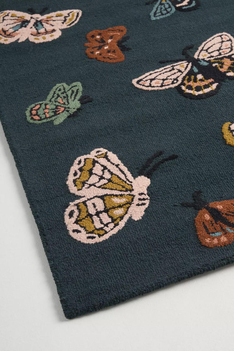 May include: A dark teal rug featuring a repeating pattern of colorful butterflies. The butterflies have detailed wing patterns in shades of pink, brown, green, and yellow. The rug has a textured, plush appearance.