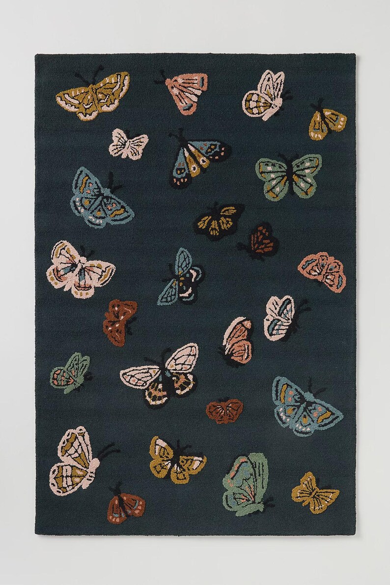May include: A rectangular rug with a dark teal background features a pattern of various colorful butterflies. The butterflies are in shades of pink, yellow, blue, and brown, with detailed wing patterns. The rug is a decorative home accent.