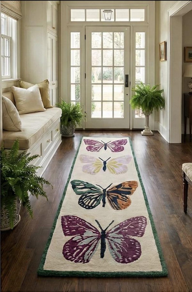 May include: A cream-coloured rectangular rug with a green border, featuring four large butterfly designs in purple, green, orange, and yellow. The rug is placed on a dark wood floor in a well-lit hallway, creating a decorative accent.