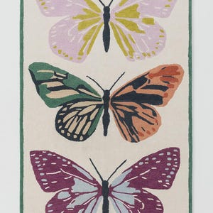 May include: A rectangular rug with three butterfly designs. The top butterfly has pink and yellow wings, the middle one has green and orange wings, and the bottom one has purple and blue wings. The rug has a cream background and a green border.