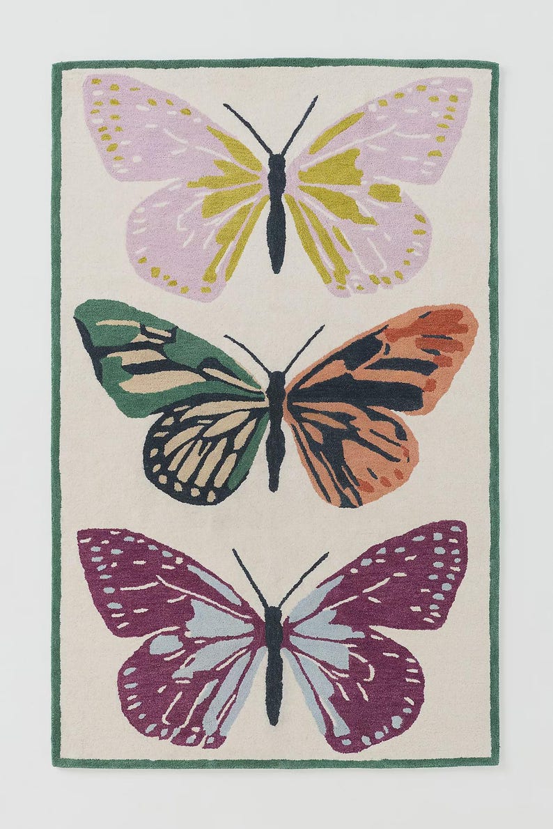 May include: A rectangular rug with three butterfly designs. The top butterfly has pink and yellow wings, the middle one has green and orange wings, and the bottom one has purple and blue wings. The rug has a cream background and a green border.