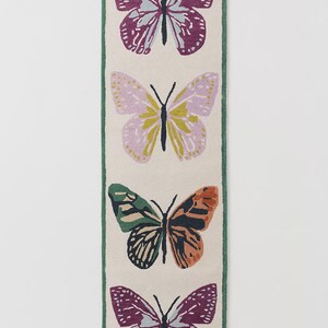 May include: A rectangular rug with four butterfly motifs. The butterflies are in shades of purple, pink, green, and orange, set against a cream background with a green border. The rug is a decorative home accent, suitable for a hallway.