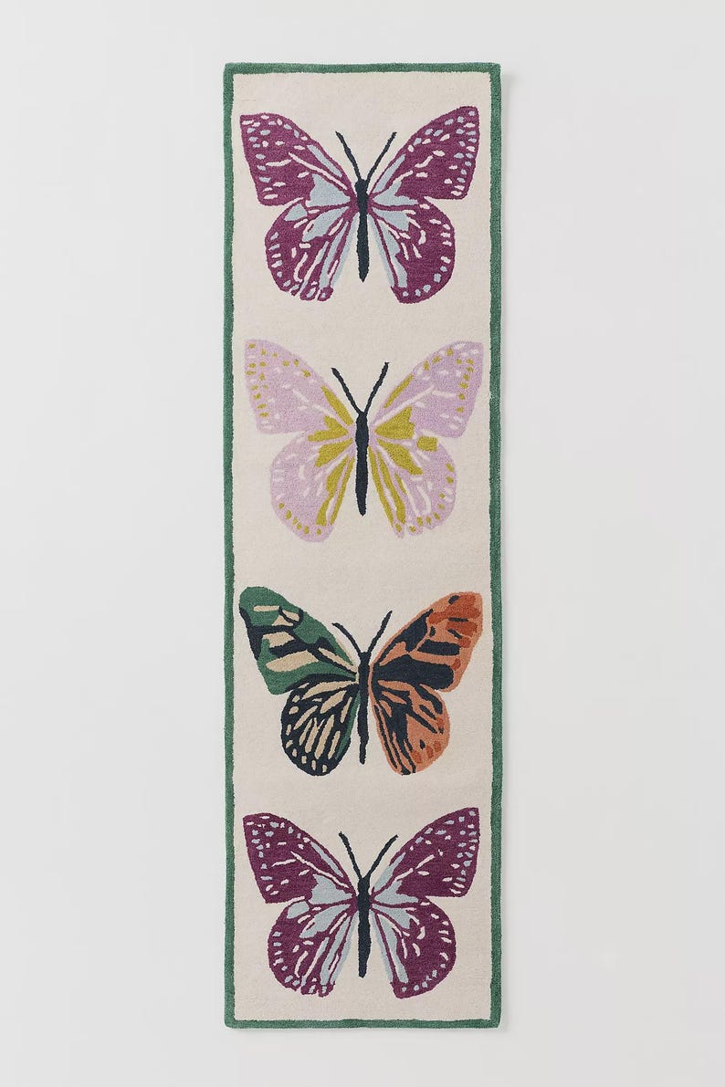 May include: A rectangular rug with four butterfly motifs. The butterflies are in shades of purple, pink, green, and orange, set against a cream background with a green border. The rug is a decorative home accent, suitable for a hallway.