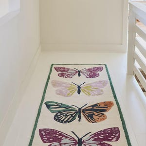 May include: A rectangular rug featuring four butterfly motifs in purple, green, orange, and yellow. The rug has a cream background and a green border. The butterflies are arranged symmetrically along the rug's length. The rug is suitable for a hallway.