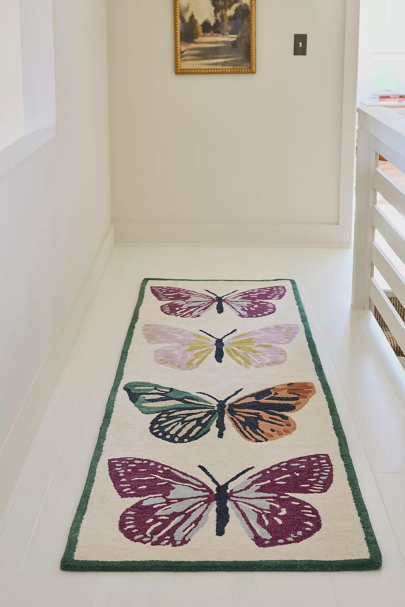 May include: A rectangular rug featuring four butterfly motifs in purple, green, orange, and yellow. The rug has a cream background and a green border. The butterflies are arranged symmetrically along the rug's length. The rug is suitable for a hallway.
