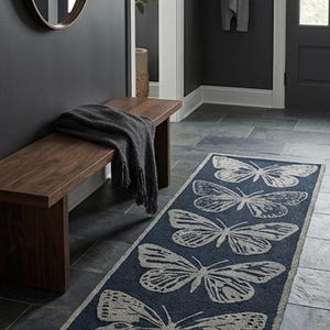 May include: A navy blue rectangular rug featuring a repeating butterfly design in light grey. The rug is laid on a dark grey tiled floor in a hallway. A wooden bench and a dark grey blanket are also visible.