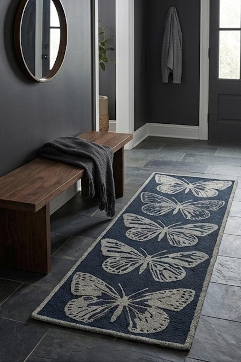 May include: A navy blue rectangular rug featuring a repeating butterfly design in light grey. The rug is laid on a dark grey tiled floor in a hallway. A wooden bench and a dark grey blanket are also visible.