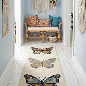 May include: A cream-coloured hallway runner rug featuring three colourful butterfly designs. The butterflies are in shades of orange, yellow, blue, and green. The rug is placed on white wooden flooring, with light blue walls and macrame wall hangings.
