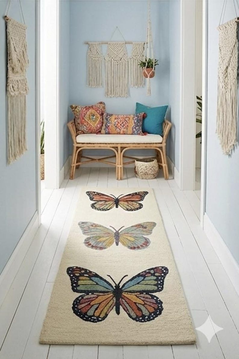 May include: A cream-coloured hallway runner rug featuring three colourful butterfly designs. The butterflies are in shades of orange, yellow, blue, and green. The rug is placed on white wooden flooring, with light blue walls and macrame wall hangings.