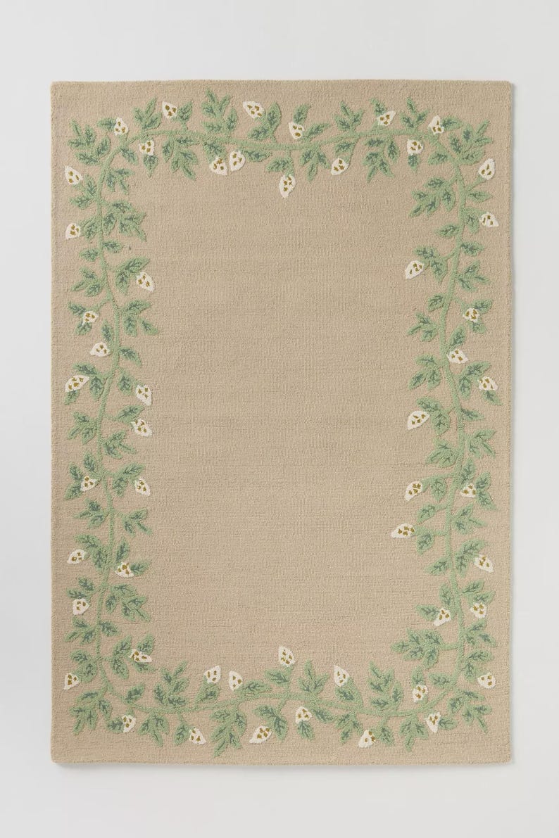 May include: Beige rectangular rug featuring an embroidered border of green leaves and white floral details. The design creates a frame around the central area of the rug, offering a decorative and natural look. Suitable for indoor use.