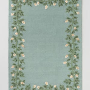 May include: A rectangular, pale teal rug with a detailed floral border. The border features green vines with dark green leaves and small, off-white flower clusters. The rug has a textured appearance.