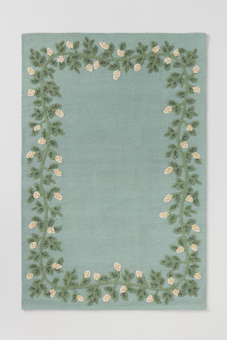 May include: A rectangular, pale teal rug with a detailed floral border. The border features green vines with dark green leaves and small, off-white flower clusters. The rug has a textured appearance.