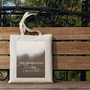 May include: A beige canvas tote bag with a rectangular image of a misty mountain landscape. The text "THE HIGHLANDS are calling, I MUST GO." is printed on the image in a serif font. The bag has two handles and is resting on a wooden bench.